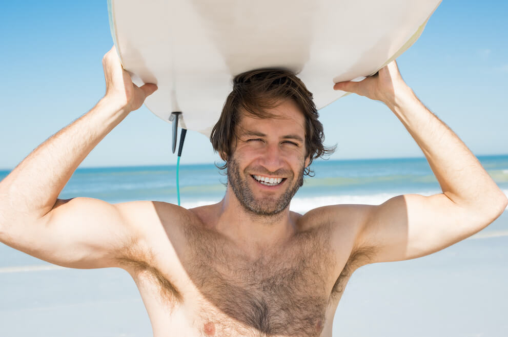 Surfer standing on beach holding board above his head.